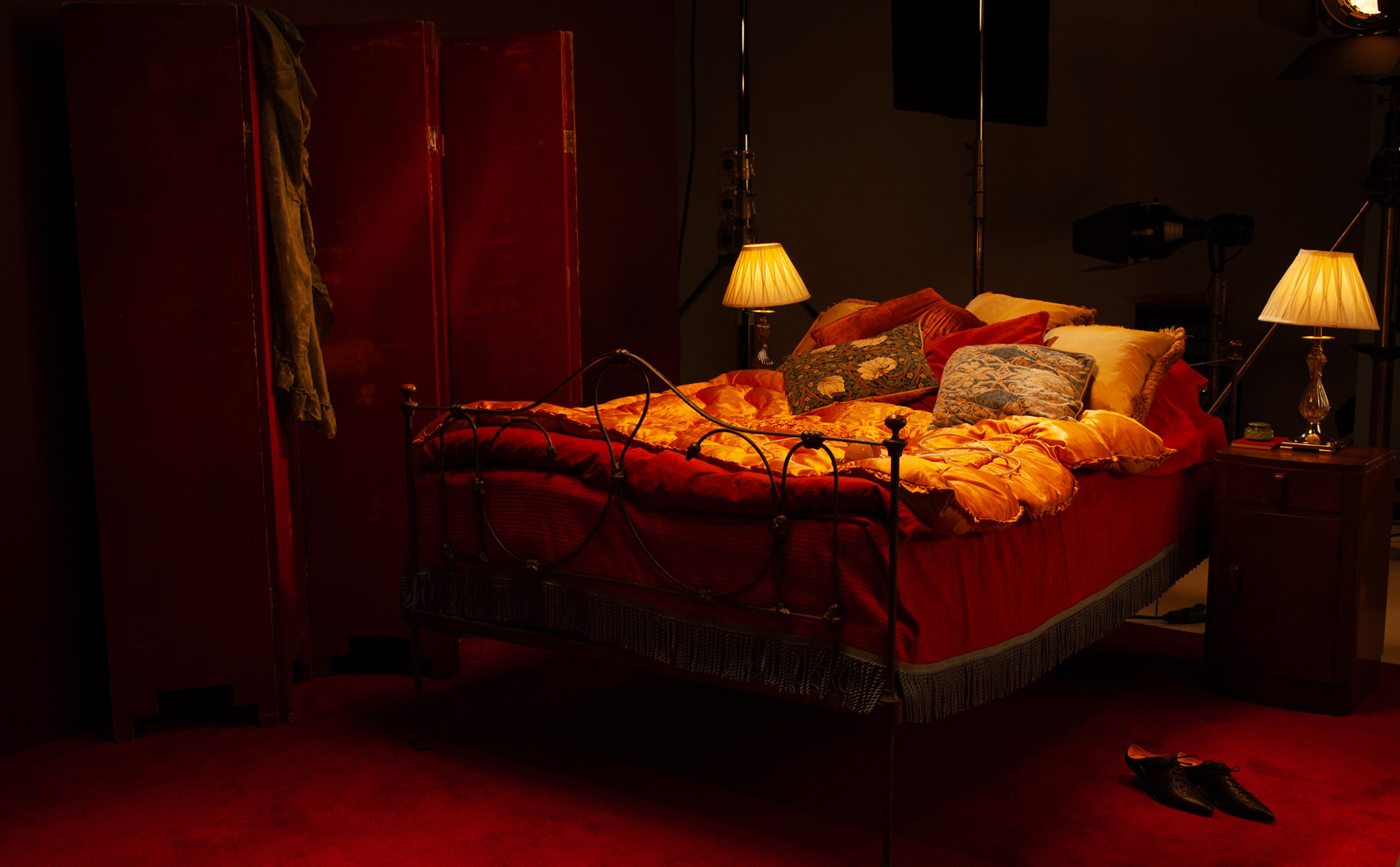 Dark bedroom with a bed covered in red blankets and yellow pillows, lit by two lamps.
